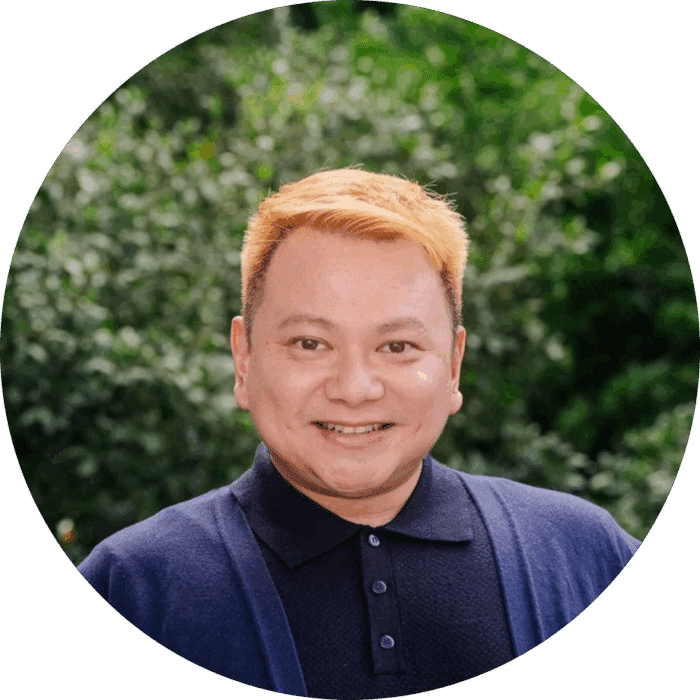 Meet the team: A person with short, light ginger hair smiles at the camera whilst wearing a dark blue collared shirt and cardigan, standing in front of a green, leafy outdoor background.