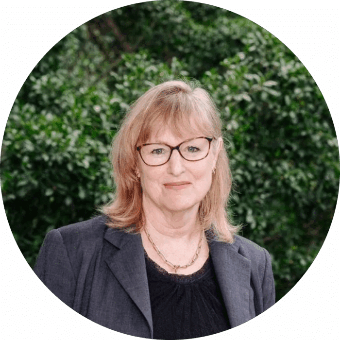 Meet the Team: A woman with light brown hair and glasses, wearing a dark blazer and black top, stands outdoors in front of green leafy bushes, smiling gently at the camera.