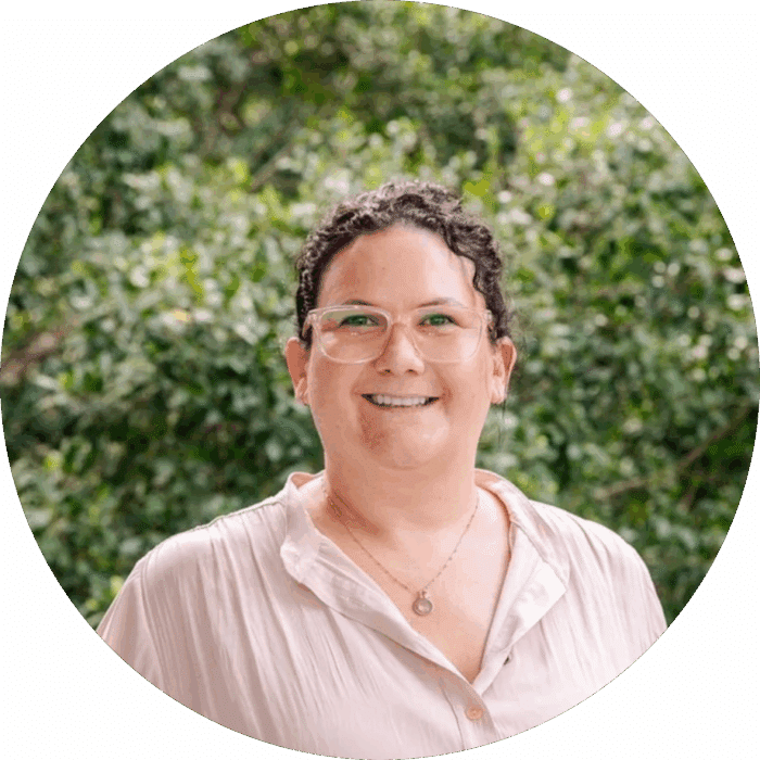 Meet The Team: A person with short, curly dark hair and glasses smiles at the camera. They are wearing a light-coloured shirt and a necklace, standing in front of a green, leafy outdoor background.