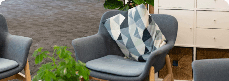 A modern grey armchair with wooden legs sits in an office space, perfect for an in-home care Sydney setting, featuring a decorative geometric-patterned cushion. A green plant and white storage cupboards are visible in the background.