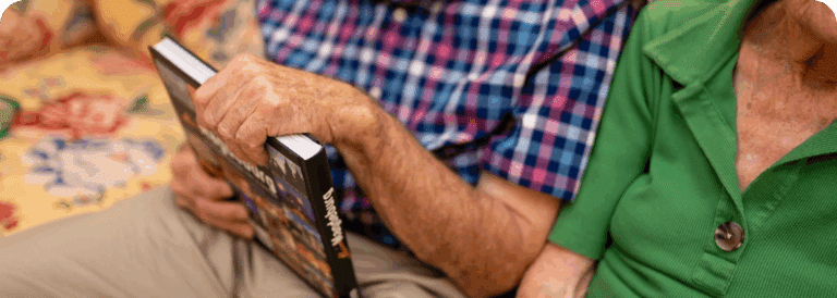 Two elderly people sit closely together on a floral-patterned sofa in a cosy living room, highlighting the warmth often found with in-home care Sydney; one holds a book in a blue and pink checked shirt, while the other wears green. Their faces are not visible.