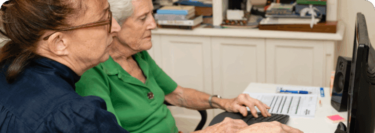 Two older women sitting at a desk, looking at a computer screen together—one types while the other observes. Papers and books fill the space, suggesting they’re exploring in-home care Sydney options.
