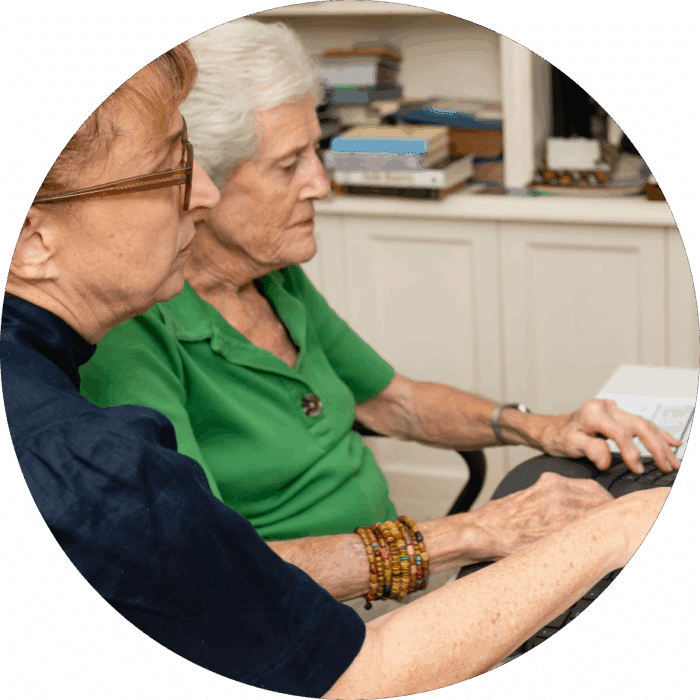 Two older women sit side by side at a desk, focusing on a computer screen. One wears glasses and a navy top, the other wears a green shirt and beaded bracelets. Books and papers are visible in the background.