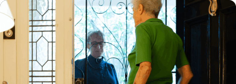 An older woman in a green shirt stands inside by an open door, warmly greeting another older person standing outside behind a decorative security screen—an everyday moment in domiciliary care Sydney.