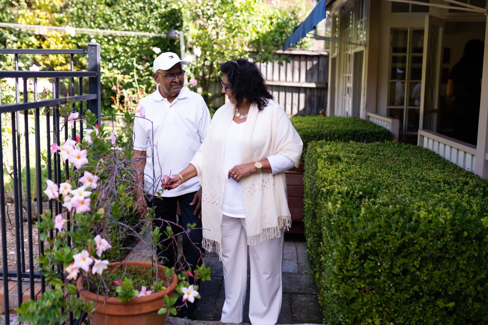 An older man and woman stand by a flowering plant in a garden, smiling and talking. The woman is touching the plant, whilst trimmed green bushes line the walkway beside their house, reflecting the joys of in-home care Sydney.