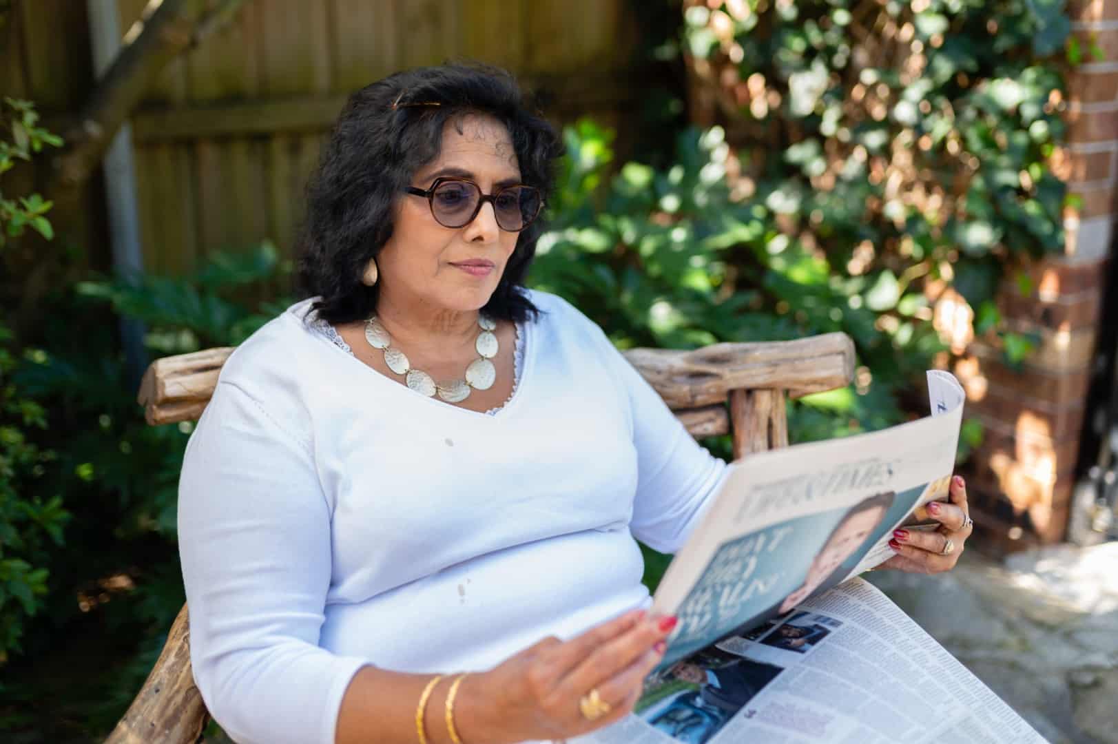 A woman with dark hair and glasses sits on a wooden bench outdoors, wearing a white top and silver necklace, reading a newspaper about in-home care Sydney. Green plants and a wooden fence are in the background.