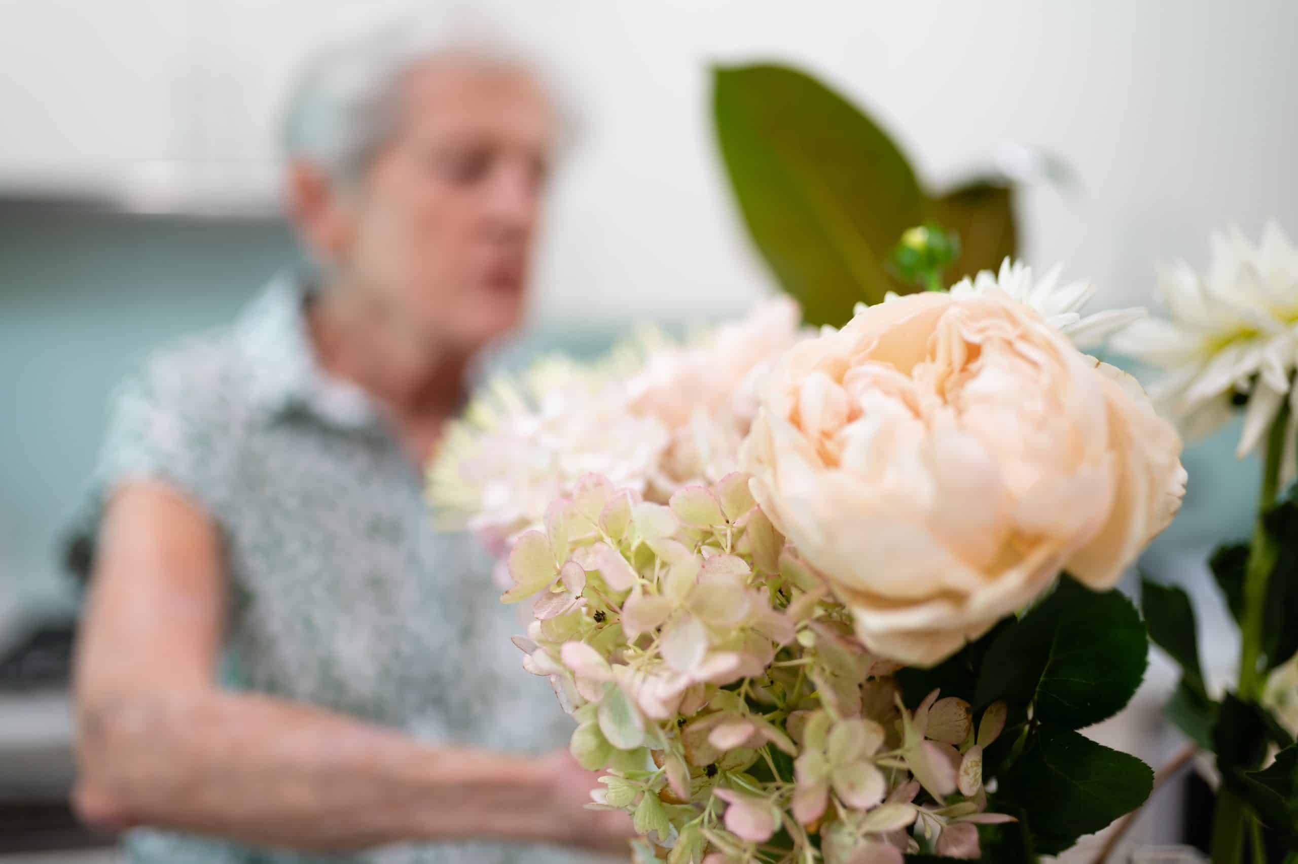 Close-up of pale pink and white flowers in sharp focus, with an older woman in a patterned shirt blurred in the background, suggesting she is arranging or admiring the bouquet as part of in-home care Sydney services.