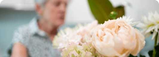 A close-up of pale pink and white flowers with an older woman blurred in the background, wearing a patterned shirt—evoking the warmth and comfort of in-home care Sydney provides.