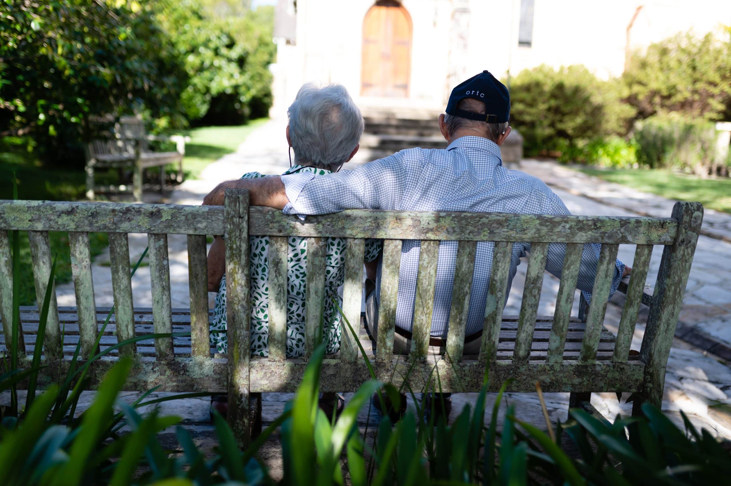 An elderly couple sit closely together on a weathered wooden bench in a garden, seen from behind. Enjoying the benefits of in-home care Sydney, they relax as sunlight filters through the trees around them.