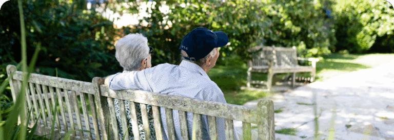 An elderly couple sit closely together on a wooden bench in a sunny park, surrounded by greenery and other empty benches, enjoying a peaceful moment—much like those cherished during quality in-home care Sydney provides.