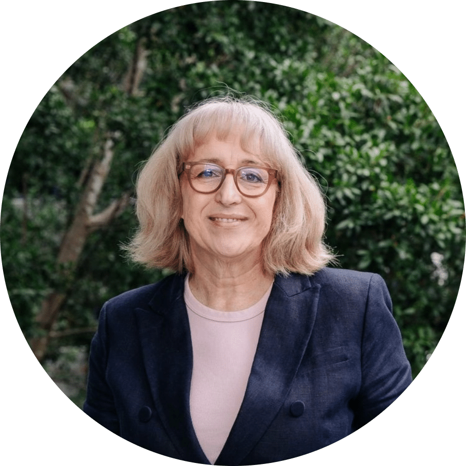 Meet The Team: A woman with light blonde hair and glasses smiles at the camera, wearing a black blazer over a pale top. She is standing outdoors in front of green foliage.