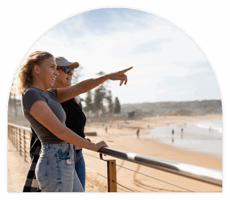 Two women standing by a seaside railing, smiling as one points towards the ocean. The sandy beach and waves are visible in the background on a bright, sunny day.