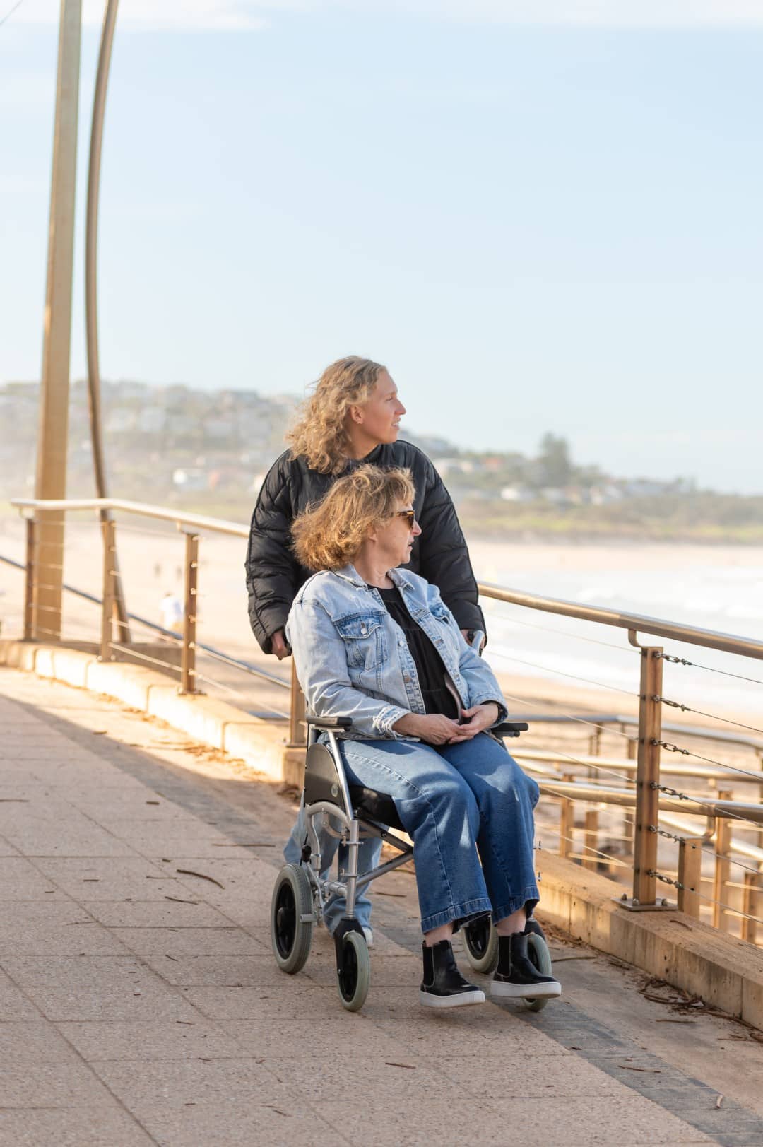A carer and a woman in a wheelchair looking out over the beach from a promenade.
