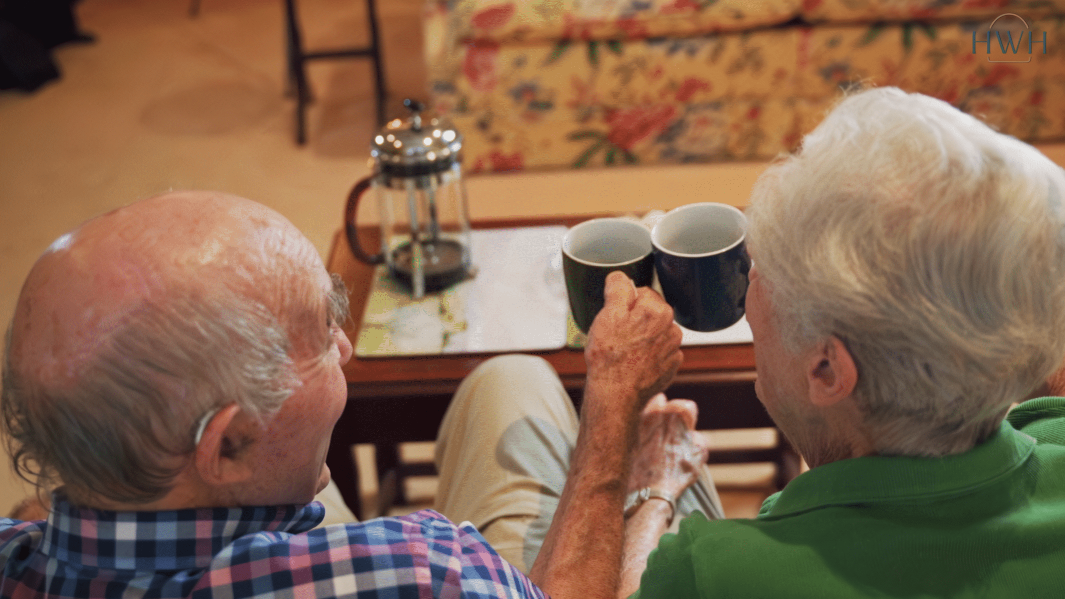 Two elderly people sit closely together on a sofa, facing away and holding coffee mugs. A cafetière rests on a small table in front of them, reflecting the warmth and comfort of in-home care Sydney provides. A floral-patterned sofa is in the background.