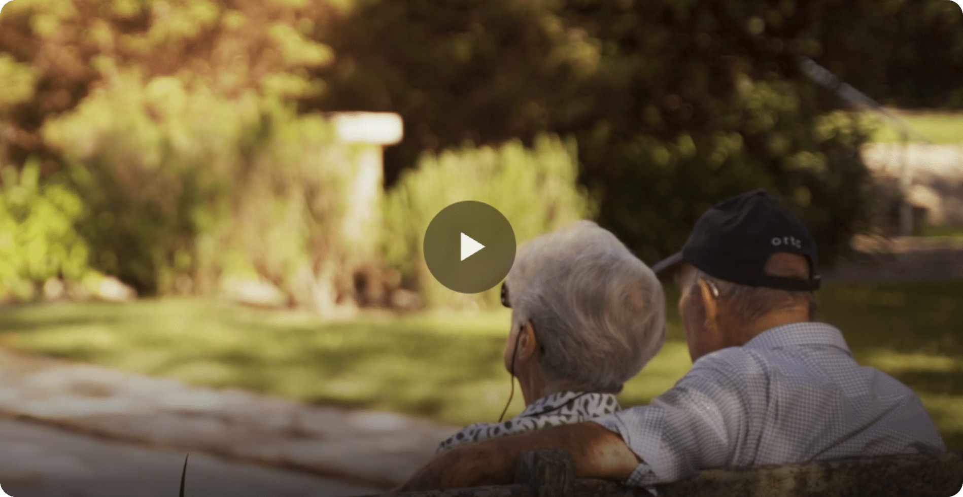 Two elderly people sit closely together on a park bench, facing away from the camera and looking at a sunlit garden. This calm, peaceful moment reflects the companionship often fostered through in-home care Sydney.