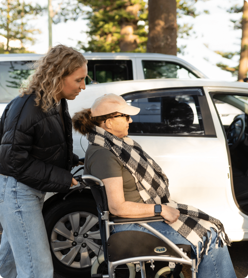 A woman with curly blonde hair, providing in-home care in Sydney, pushes another woman in a wheelchair beside a parked white car. The seated woman wears a hat, sunglasses, and checked scarf. Trees and parked vehicles are visible in the background.
