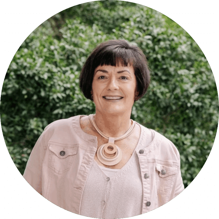 Meet The Team: A woman with short dark hair smiles outdoors, wearing a pale pink jacket, a matching top, and a chunky circular necklace. Lush green foliage is visible in the background.