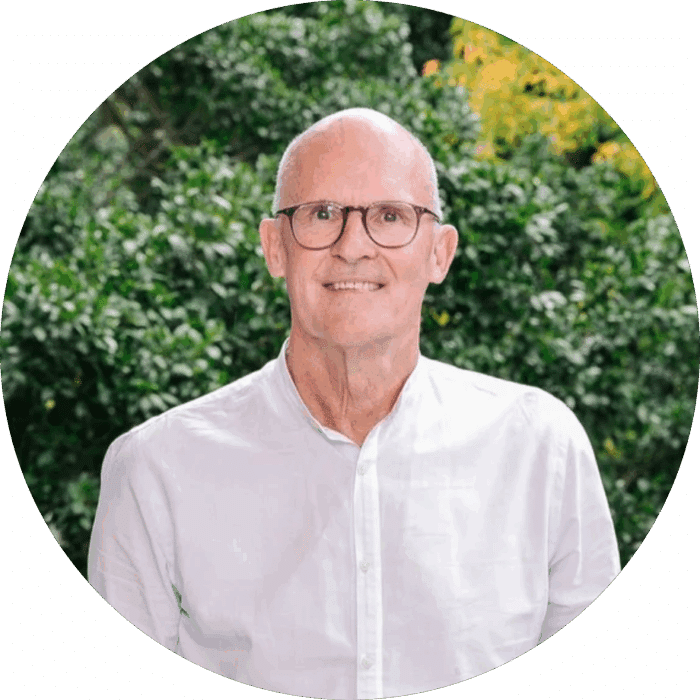 Meet The Team: A smiling older man with glasses and a white button-up shirt stands outdoors in front of green foliage.
