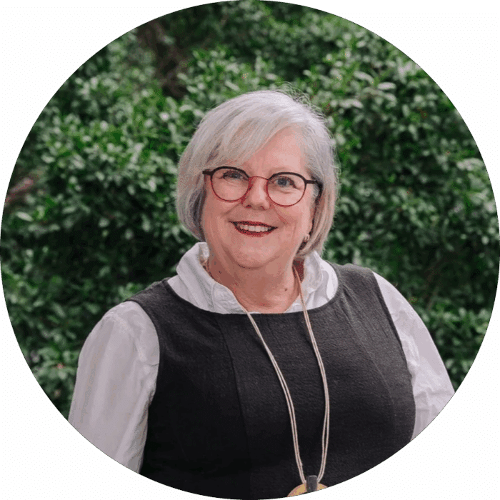 Meet The Team: Smiling older woman with short grey hair, glasses, and red lipstick, wearing a white blouse and black waistcoat, standing outdoors in front of green foliage.