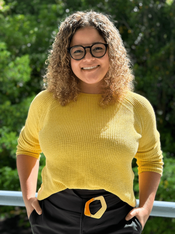 Meet The Team: A woman with curly hair and glasses smiles outdoors, wearing a yellow jumper and black trousers with a yellow-buckled belt. Green foliage is visible in the background.