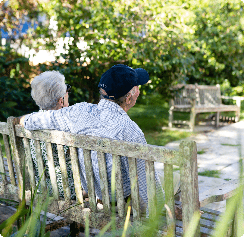 An elderly couple sit closely together on a wooden bench in a sunny park, surrounded by greenery. The man has his arm round the woman, and they are both facing away, enjoying the peaceful outdoor setting.