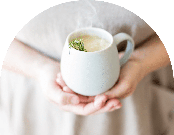 A person holding a steaming white mug filled with a hot beverage, garnished with a sprig of rosemary. The background is soft and out of focus, highlighting the cup and hands.