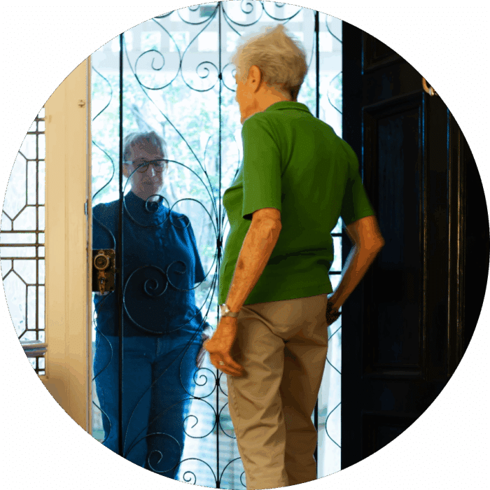 An older adult in a green shirt stands inside by a decorative metal screen door, facing a visitor outside who is wearing glasses and a dark jacket.