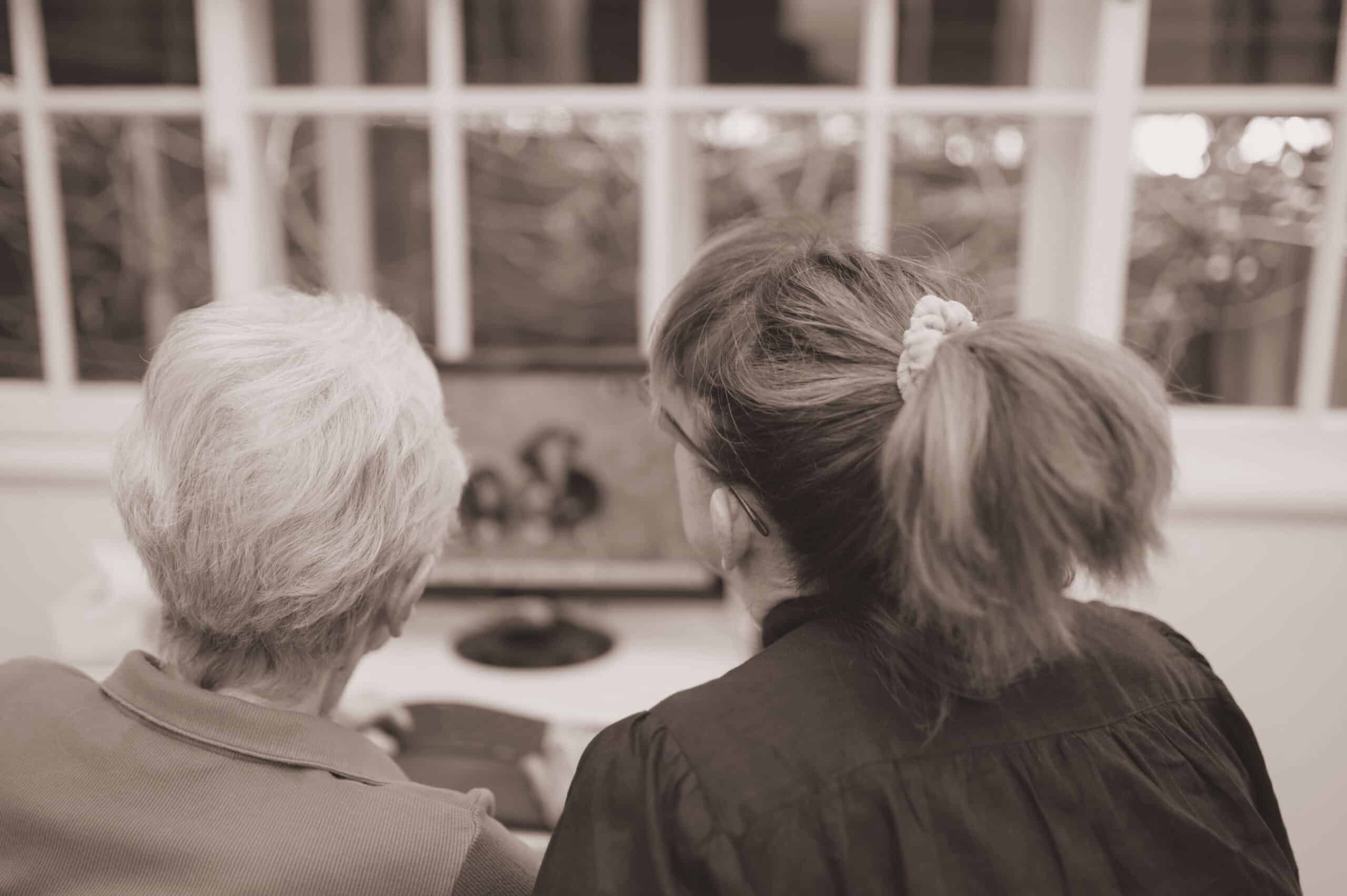 An older woman and a carer sitting side by side at home, looking out through a window together.