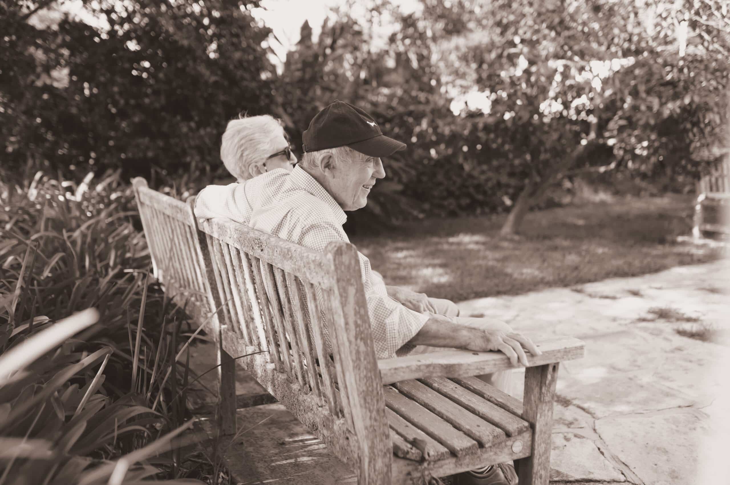An older couple sitting close together on a wooden bench in a sunlit garden.