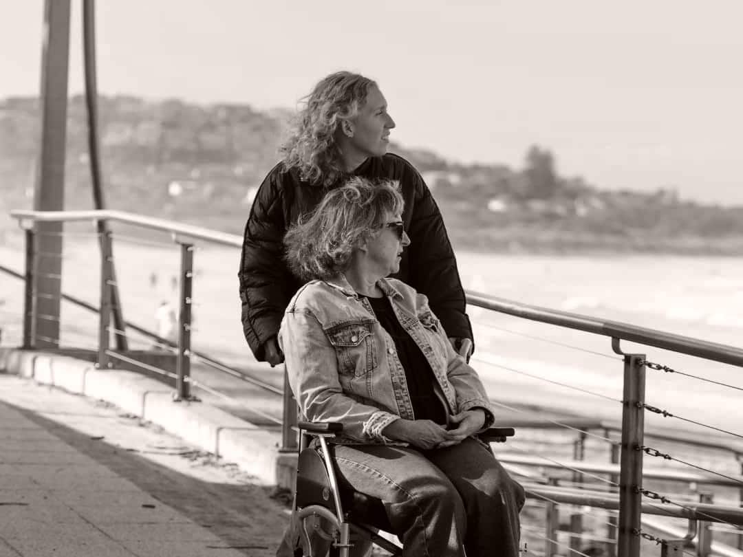 A carer standing beside a woman in a wheelchair, looking out over the beach from a promenade