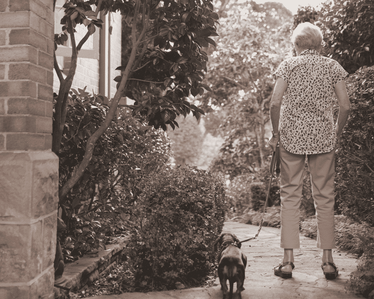 An older woman walking her dog along the garden path at her home.